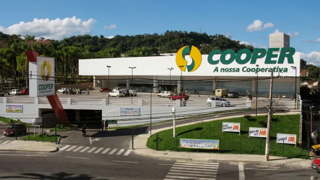 Descrição da imagem: Fachada do supermercado Cooper, no Bairro Água Verde em Blumenau, com o grande letreiro amarelo, verde e branco no alto da construção.
A fotografia mostra a vista frontal de um supermercado de grande porte, com fachada branca e estrutura moderna. No alto do prédio, à direita, está o logotipo da marca "COOPER", com um símbolo circular nas cores amarela e verde, e ao lado o texto em letras verdes "COOPER" e, abaixo, "A nossa Cooperativa". À esquerda da imagem, há uma placa vertical, também branca, repetindo o mesmo logotipo e nome da loja, fixada em uma estrutura triangular vermelha que se conecta ao prédio principal.
Em frente ao supermercado há um grande estacionamento pavimentado, com diversos carros de cores variadas estacionados, entre eles tons de branco, prata, vermelho e azul. Algumas pessoas caminham próximas às entradas e à área de carrinhos de compras, visível à direita da imagem. A entrada de vidro do supermercado fica no centro da fachada, com portas amplas e transparentes.
Abaixo do estacionamento, há uma área com paredes claras onde se lê a palavra "FARMÁCIA" em uma placa, indicando uma farmácia integrada ao espaço. Um pouco mais à esquerda, há um cartaz colorido de propaganda de algum produto.
Na parte inferior da imagem, um canteiro gramado leva a uma rotatória e a um cruzamento de ruas, com faixas de pedestres pintadas em branco. Sobre o gramado, há três placas de propaganda com preços promocionais em vermelho, como "198", "148" e "138", além de uma faixa horizontal com informações sobre limpeza. Um poste de iluminação e fios elétricos aparecem à direita, típicos de uma via urbana.
Ao fundo da imagem, à esquerda, vê-se um morro coberto por vegetação densa, com árvores altas, e o céu está azul claro com algumas nuvens brancas espalhadas, sugerindo um dia ensolarado. À direita ao fundo, aparecem outras construções residenciais, reforçando o contexto urbano do bairro.