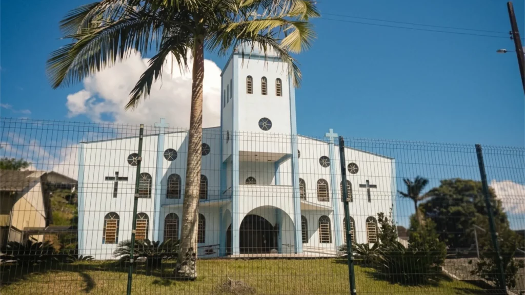 Descrição da imagem: Fachada da Paróquia Nossa Senhora do Perpétuo Socorro, fotografada em dia ensolarado, com uma grande palmeira em primeiro plano.
A fotografia mostra a frente de uma igreja de arquitetura moderna, pintada em branco com detalhes em azul-claro nas bordas e cantos das paredes. A construção tem dois níveis de altura irregular: à esquerda, um corpo mais baixo com telhado inclinado, e à direita, uma torre alta e estreita que se destaca no centro da fachada, funcionando como campanário. No topo dessa torre há três pequenas janelas retangulares em arco, e um pouco abaixo, uma janela circular em formato de roseta.
Na fachada há diversas cruzes cristãs pintadas em preto, distribuídas simetricamente nos dois lados da entrada, além de várias janelas em formato de arco com esquadrias em tom de madeira. No centro da construção, um grande arco escuro marca a entrada principal da igreja, ladeado por pequenas plantas do tipo cica.
Em primeiro plano, uma alta palmeira imperial cresce bem no meio da cena, com seu tronco claro e liso e as folhas verdes se espalhando na parte superior da imagem, quase tocando o topo da torre. À frente de toda a construção há uma cerca alta de tela metálica (alambrado), sustentada por postes verdes, que separa o terreno da rua.
Ao fundo, à esquerda, aparecem outras casas e um morro com vegetação, e à direita da imagem há árvores e mais construções residenciais ao longe. O céu está azul e claro, com uma nuvem branca visível atrás da torre da igreja, reforçando o clima ensolarado do dia em que a foto foi tirada.