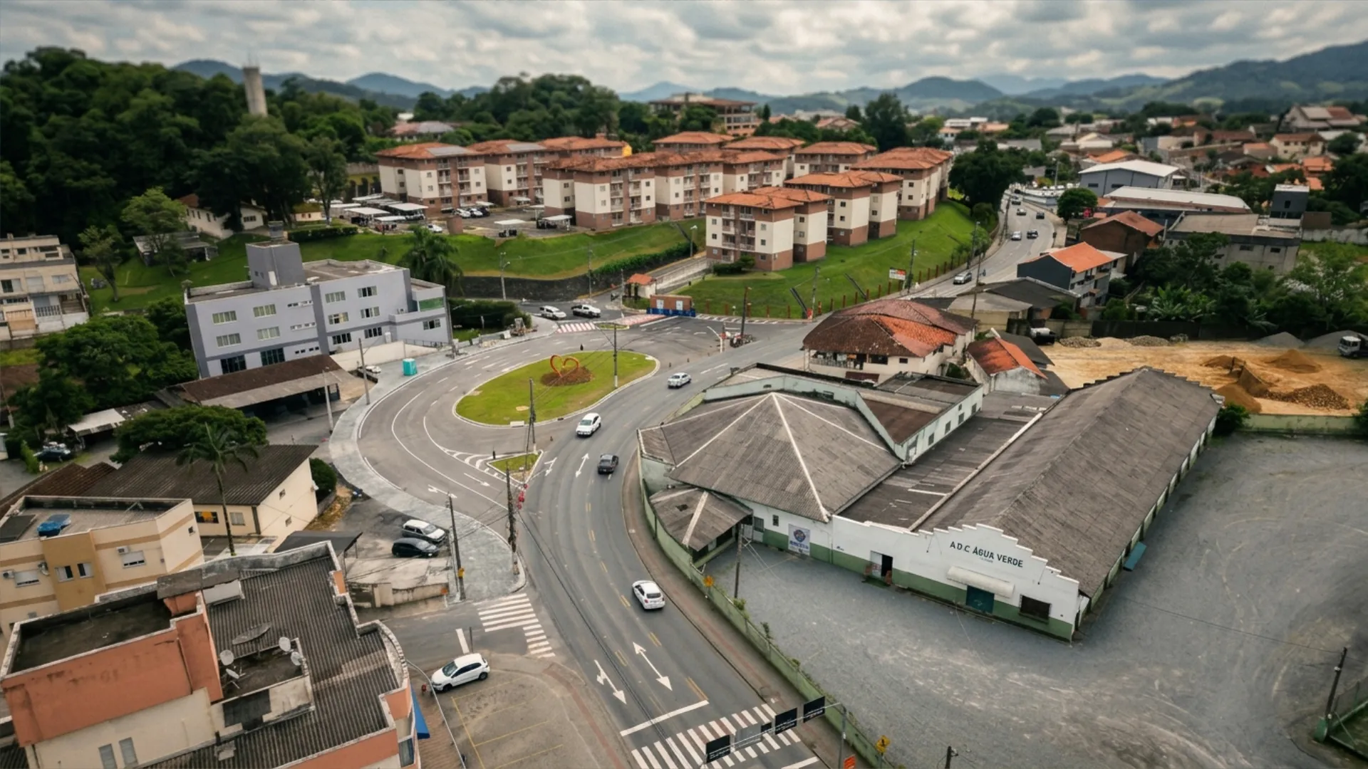 Descrição da imagem: Vista aérea do Bairro Água Verde, em Blumenau, mostrando uma rotatória de trânsito no centro da cena. A fotografia foi tirada de cima, provavelmente por um drone, em um dia claro. No centro da imagem está uma rotatória viária com um pequeno canteiro gramado oval em seu meio. Diversas ruas se encontram nesse ponto, formando o cruzamento, com faixas de pedestres pintadas em branco e amarelo próximas ao acesso das vias. Alguns carros brancos e escuros circulam pela rotatória e pelas ruas ao redor. À esquerda e ao fundo da imagem, no topo, aparece um conjunto de prédios residenciais de médio porte, com fachadas claras e telhados em tom terracota, dispostos lado a lado sobre uma área gramada. Atrás desses prédios, há uma extensa vegetação verde e densa, sugerindo uma área de mata ou morro. À direita da rotatória, destaca-se um grande galpão com telhado escuro e formato triangular bem inclinado, com paredes de tijolo aparente e pintura clara na parte inferior. Ao lado desse galpão há um pátio de terra clara. Na parte inferior da imagem, veem-se casas menores, de um a dois pavimentos, com telhados variados em tons de vermelho e cinza, típicas de bairro residencial, além de alguns quintais, muros e veículos estacionados nas ruas. No canto superior direito, é possível notar mais construções e ruas que se estendem para outras partes do bairro, reforçando o caráter urbano e conectado da região. De modo geral, a imagem transmite um retrato típico da paisagem urbana de Blumenau: uma mistura de área residencial com prédios e casas, comércio local (representado pelo galpão), infraestrutura viária organizada em torno da rotatória, e a presença constante da natureza ao fundo, com morros cobertos de vegetação característicos da região.