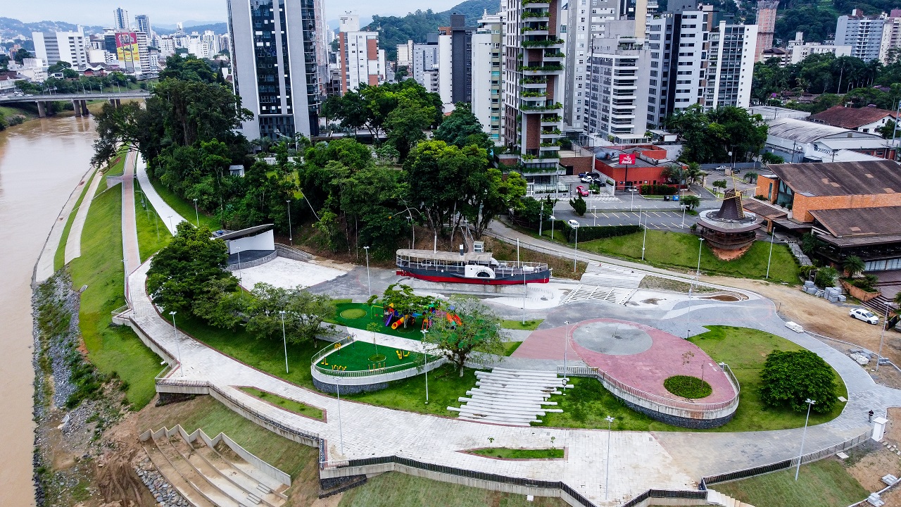 Esta vista aérea impressionante captura a essência da Praça Juscelino Kubitschek de Oliveira, carinhosamente chamada pelos locais de 'Prainha', localizada na margem esquerda do imponente Rio Itajaí-Açu, no bairro Ponta Aguda em Blumenau, Santa Catarina. A imagem destaca a infraestrutura moderna resultante de uma recente e aclamada revitalização. O calçadão sinuoso de concreto convida para caminhadas e passeios de bicicleta, conectando as diferentes áreas de lazer da praça. Um dos destaques centrais é a embarcação turística ancorada, o Barco do Parque, que se tornou um símbolo icônico do local, oferecendo um vislumbre da história fluvial da cidade. A foto revela ainda o vibrante playground infantil colorido, pronto para a diversão das crianças. Próximo a ele, uma praça de eventos circular com degraus em anfiteatro, ideal para apresentações culturais e encontros comunitários. A vegetação densa ao redor da praça contrasta belamente com o concreto, criando um oásis verdejante no coração urbano. As escadas e rampas dão acesso direto à margem do rio, permitindo uma conexão mais próxima com a natureza. Ao fundo, o horizonte de Blumenau se estende, com edifícios residenciais e comerciais imponentes no centro da cidade, incluindo a famosa 'Curva do Rio'. Estruturas viárias podem ser discernidas ao longe, mostrando a integração da praça com a malha urbana. Esta foto encapsula o charme de Blumenau: a combinação de história, natureza e modernidade. A Prainha na Ponta Aguda é um ponto de encontro essencial para moradores e um destino imperdível para turistas que desejam relaxar e apreciar a beleza do Vale do Itajaí.