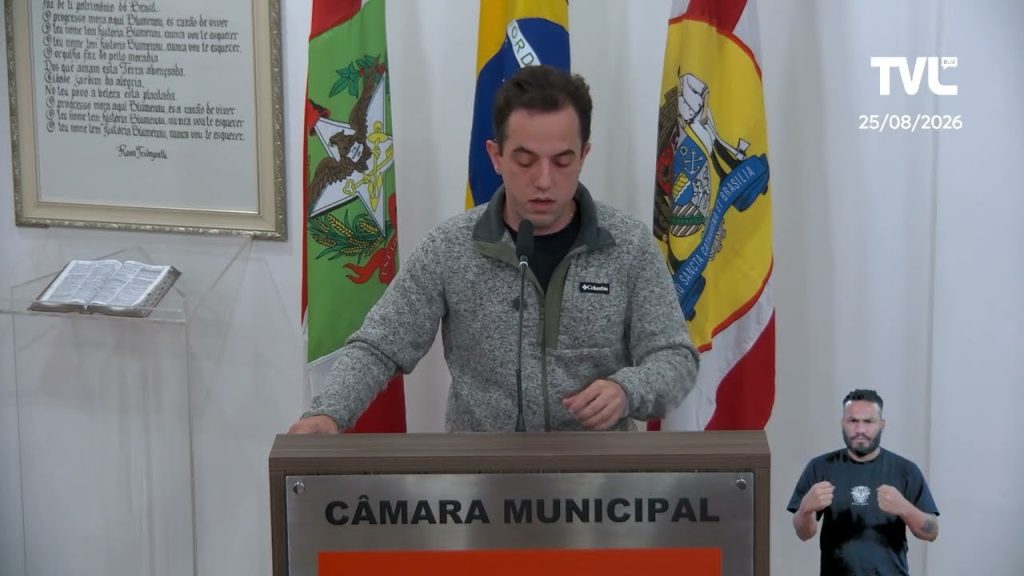 A imagem mostra o vereador Flávio Linhares discursando na tribuna da Câmara Municipal de Blumenau, transmitido pela TVL Blu no dia 25/08/2026. Ele está de pé diante do púlpito, vestindo um casaco cinza de lã com detalhes em verde musgo por baixo, enquanto lê seu pronunciamento em um microfone fixado à tribuna, que traz a inscrição "Câmara Municipal" em destaque. Ao fundo, três bandeiras estão dispostas lado a lado: à esquerda a bandeira de Blumenau, ao centro a bandeira do Brasil, e à direita a bandeira de Santa Catarina, todas emolduradas por um painel branco. No canto superior esquerdo da cena, aparece um quadro emoldurado com um poema dedicado à cidade de Blumenau, e logo abaixo dele, sobre um suporte de acrílico, está exposta uma Bíblia aberta. No canto inferior direito da imagem, um intérprete de Libras realiza a tradução simultânea do discurso, garantindo acessibilidade à transmissão. No canto superior direito, consta o logotipo da emissora TVL Blu junto com a data da sessão.